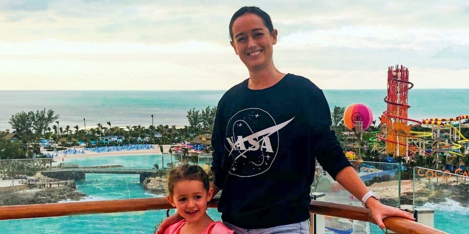 Cruise Mummy and her young daughter pose on a balcony at CocoCay, with the ocean and water park attractions in the background. The mother is wearing a NASA sweatshirt and the daughter is smiling, both enjoying their day at the island destination.