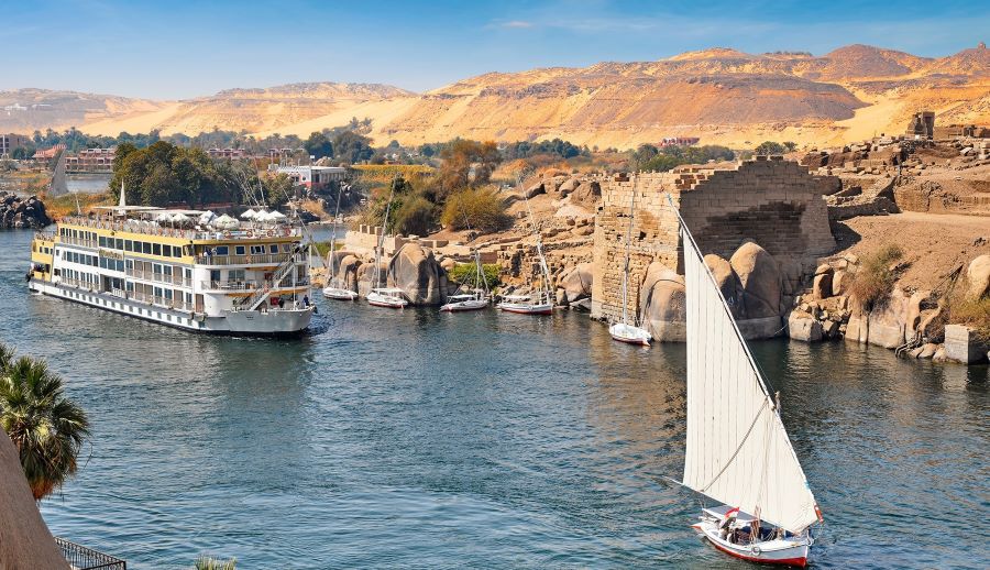 The AmaDahlia, AmaWaterways' luxury river cruise ship, navigates the historic Nile River. A traditional sailboat with a single white sail glides in the foreground, contrasting with the grandeur of the cruise ship. In the background, the rugged hills of Egypt's landscape and remnants of ancient structures blend the past and present, capturing the essence of cruising the Nile.