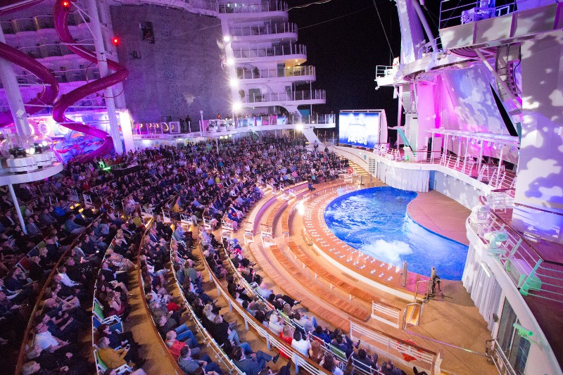 The AquaTheater on Symphony of the Seas, filled with an audience watching a nighttime performance. The theater features tiered seating surrounding a central pool used for water shows, with vibrant lighting and stage effects. A large water slide is visible on the left, adding to the dynamic and exciting atmosphere of the venue.