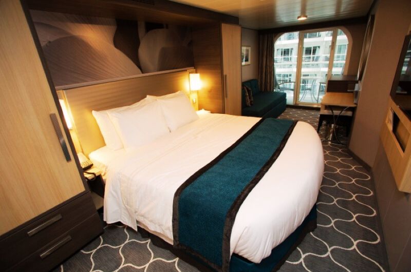 The cozy Boardwalk View Stateroom with a Balcony onboard Harmony of the Seas, featuring a plush queen-sized bed with a teal runner, a sitting area, and a balcony overlooking the bustling boardwalk area of the ship.