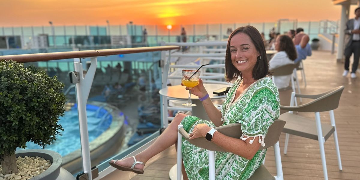 Cruise Mummy in a green patterned dress is seated on a cruise ship's deck, smiling at the camera while holding a tropical drink. The sun is setting in the background, casting a warm glow over the ocean and the ship's pool area, where other passengers are relaxing. The scene captures a moment of leisure and enjoyment during a sunset at sea.