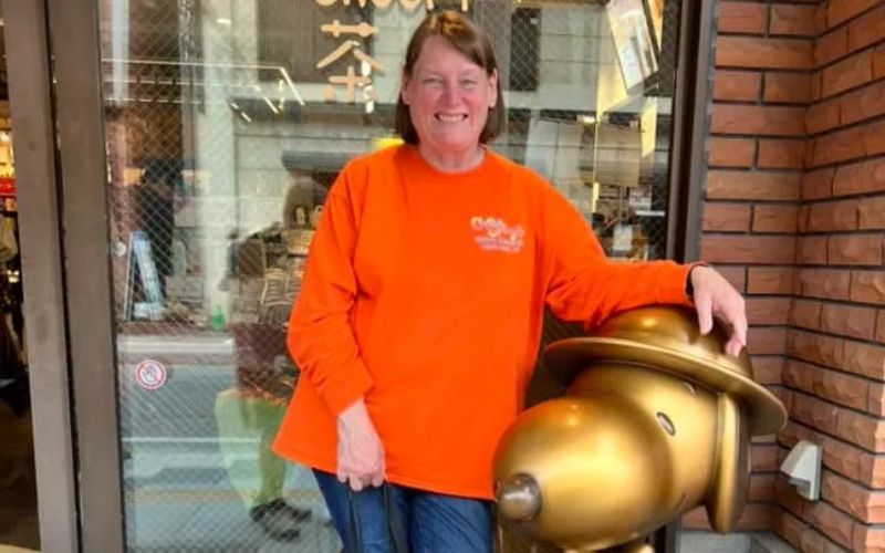 Hefty Bills smiling in front of a storefront next to a whimsical golden statue, her bright orange long-sleeve shirt adding a pop of color to the cheerful urban scene.