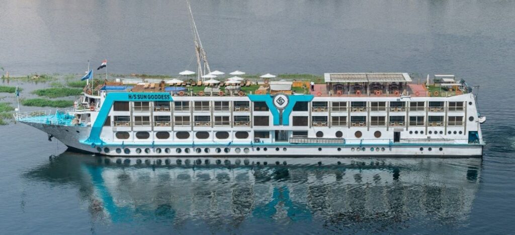 The MS Sun Goddess, a multideck river cruise ship, majestically floats on the Nile River. Its sleek white structure with bold turquoise accents is marked by the sun emblem, symbolizing the vessel's name. Passengers can be seen enjoying the sundeck loungers, promising a relaxing journey with scenic river views and Egyptian hospitality.