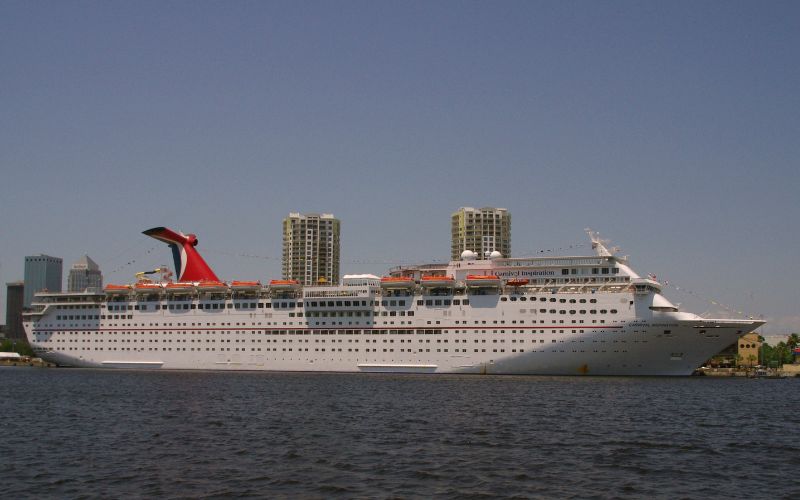 A large white cruise ship with a distinctive red funnel docked in a harbor, with modern high-rise buildings in the background under a clear blue sky, evoking a sense of unexpected travel issues with the mention of insurance.