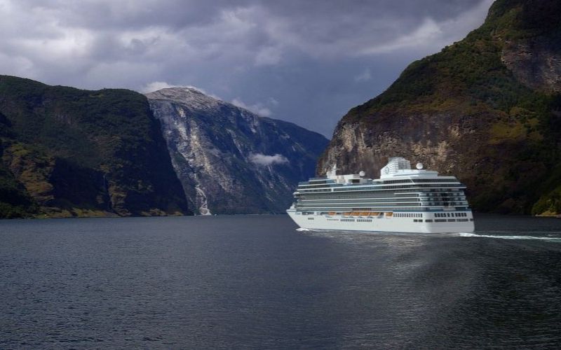 A large white cruise ship named "Oceania Vista" sails through a narrow fjord surrounded by steep, forested cliffs and a cloudy sky. Snow-capped mountains are visible in the background, creating a dramatic and serene landscape.