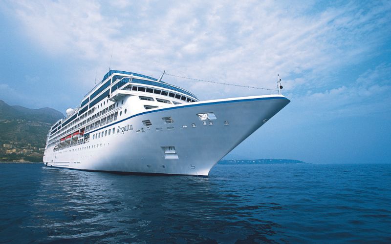 A close-up view of the cruise ship "Regatta" sailing on a calm sea. The ship's sleek white hull contrasts against the blue water and sky, with distant mountains visible in the background. The name "Regatta" is clearly displayed on the side of the ship.