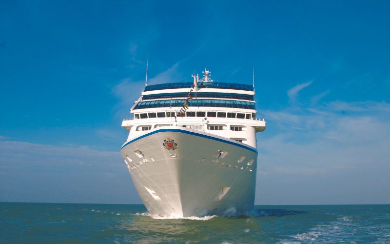 The image shows the front view Oceania Sirena cruise ship sailing on the open sea under a clear blue sky. The ship is white with multiple decks and windows, and a colorful crest is visible on the bow.