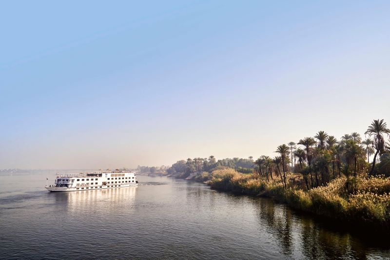 A TUI River Cruise ship smoothly sails the Nile, captured in the soft light of the morning. The ship's white exterior with rows of windows reflects on the water, contrasting with the lush greenery and palm trees on the riverbank. The tranquil waters and clear skies create a serene and picturesque scene of river cruising in Egypt.