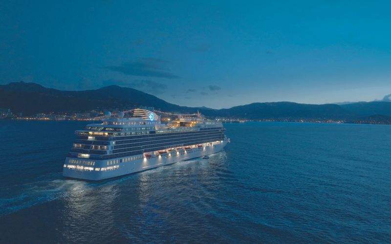 A large cruise ship, the "Allura," illuminated at night, sails away from a coastal city. The ship's lights reflect on the calm waters as the silhouette of mountains frames the horizon under a deep blue sky.