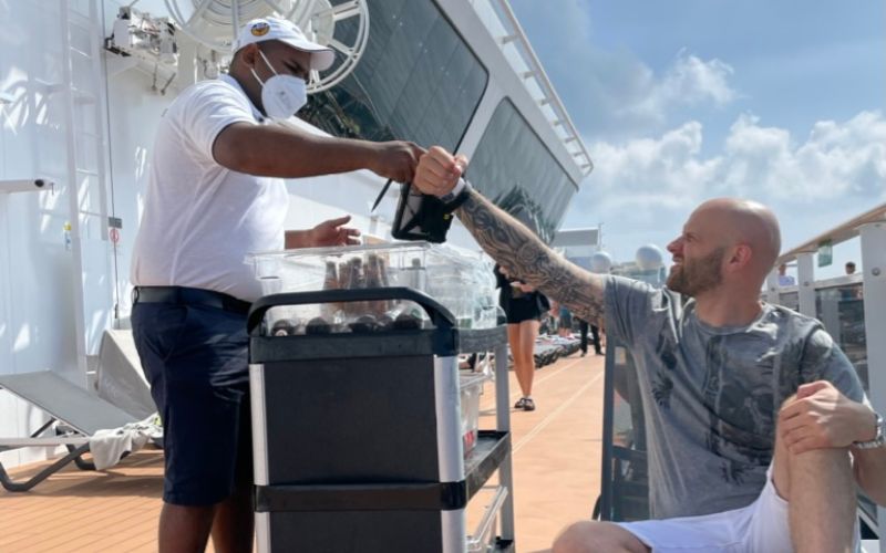 A crew member in a white shirt hands over a beverage tomy husband seated on the sunny deck of the MSC Virtuosa cruise ship.