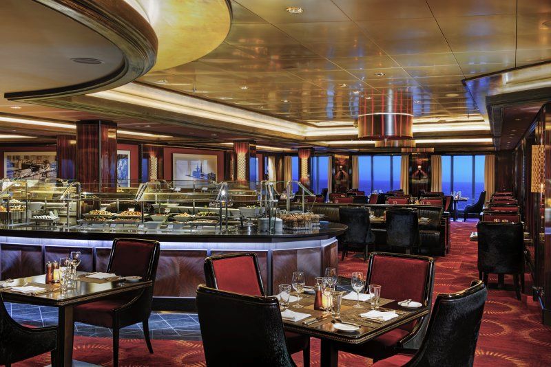 The dining area of Moderno on the Norwegian Epic cruise ship, showcasing an upscale buffet with a variety of dishes. The room is decorated with rich, dark wood tones, plush red and black seating, and elegant lighting. Tables are set for dining with a mix of ambient and overhead lighting creating a cozy atmosphere. Large windows in the background offer views of the ocean.