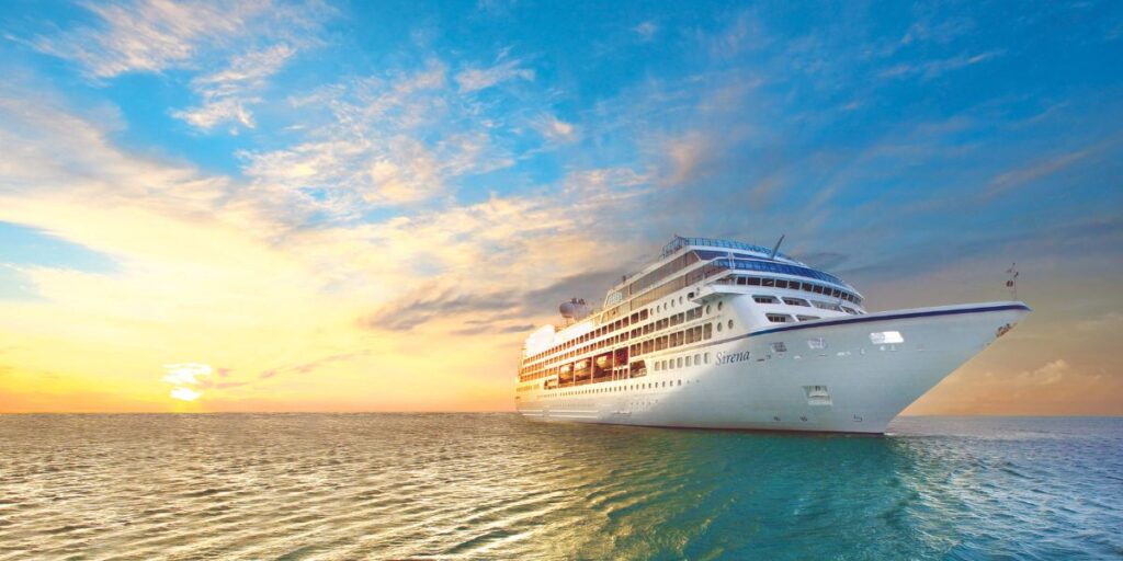 A large white cruise ship named "Sirena" sails on calm waters during a picturesque sunset, with a vibrant sky blending shades of blue, orange, and yellow.